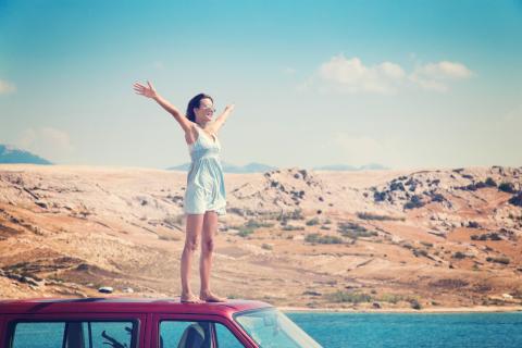 Lifestyle: Beautiful tanned woman in a blue dress standing barefoot on roof of red van spreading arms into the wind, island of Pag in Croatia.