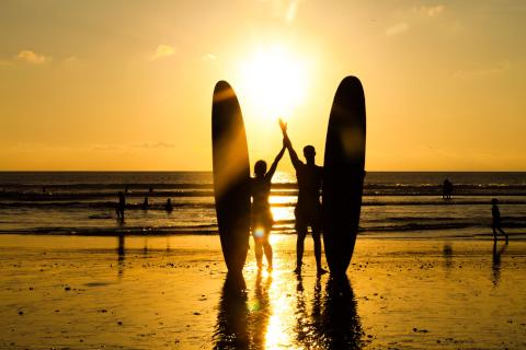 Lifestyle: Couple surfers standing on beach at sunset with their surf boards in silhouette, Bali.