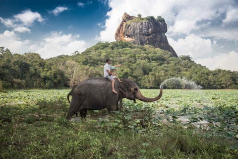 Travel: Man and child riding on the back of elephant with rock of Sigiriya as backdrop, Sri Lanka.